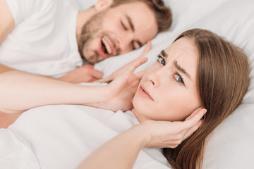 dissatisfied woman plugging ears with hands and looking at camera while lying in bed near snoring husband