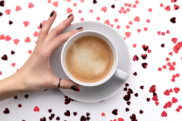 Red sparkles confetti in heart shape and cup of coffee with womans hand on white background