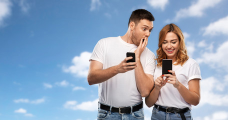 technology and people concept - happy couple in white t-shirts with smartphones over blue sky and clouds background