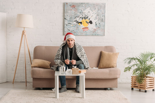 sick man in santa hat, wrapped in blanket, sitting on sofa near table with medicines