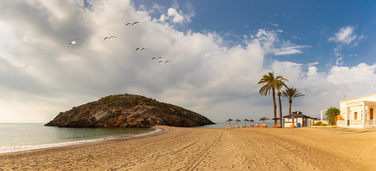 A beautiful beach in Mazarron in Spain with a big rock in the background. On the beach are palm...