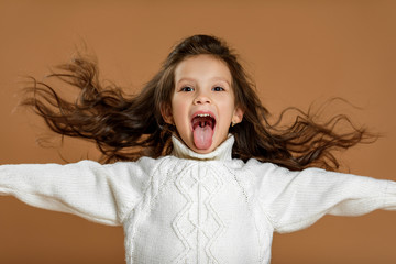 happy funny little child girl in white sweater showing her tongue on beige background. facial...
