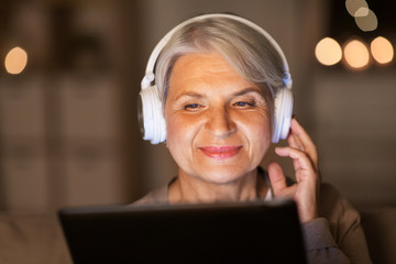 technology, people and lifestyle concept - happy senior woman in headphones and tablet pc computer listening to music at home in evening