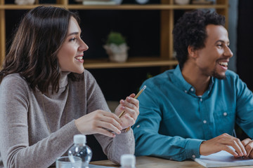 young businesswoman holding pen while sitting near african american colleague