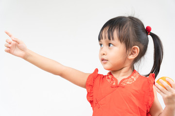Adorable asian little girl with beautiful eyes in the red chinese dress is holding the orange and she is pointing on the right and smiling with fully happiness moment.