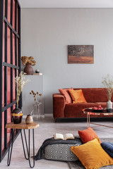 Real photo of a red couch in bright living room interior with a painting on white wall and a book on a pouf