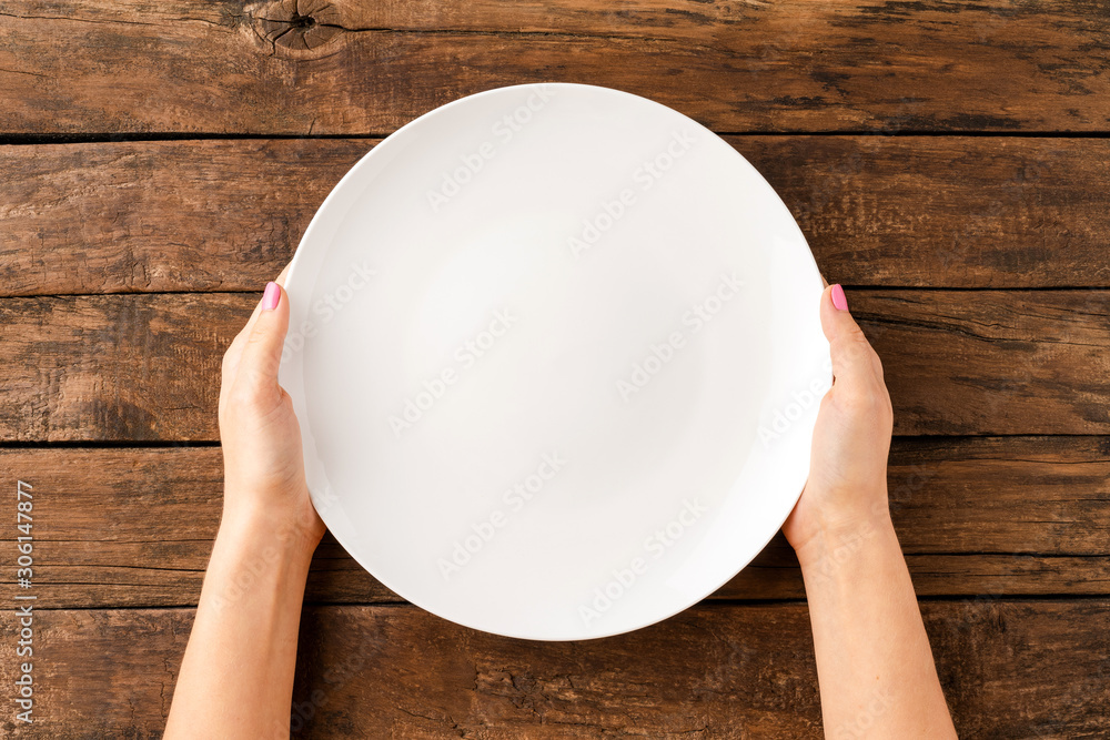 Wall mural Woman’s hands holding empty white plate over rustic wooden table. Top view