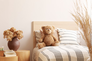 Flowers in brown vase on wooden nightstand table next to single bed with stripped bedding with teddy bear