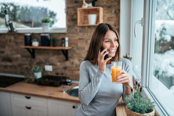 Young smiling woman talking on mobile phone, holding glass of orange juice in the kitchen.