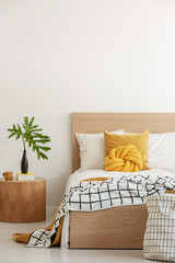 Green leaf in black vase on wooden nightstand next to Yellow pillows and blanket on white single wooden bed