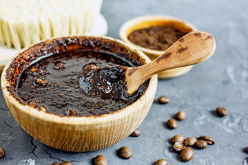 Coffee scrub in a wooden bowl with a wooden spoon on a dark stone background. Nearby is a bowl with coffee grounds, coffee grains. Coffee grounds body scrub. Close-up of mine space.