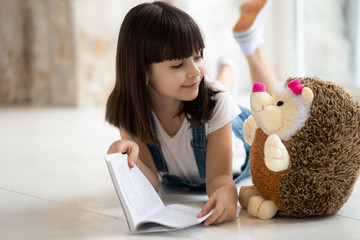 Cute small girl reading book to toy hedgehog