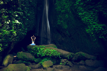 Woman sitting on the rock, practicing yoga. Young woman raising arms with namaste mudra near waterfall. Leke Leke waterfall, Bali. View from back. - obrazy, fototapety, plakaty