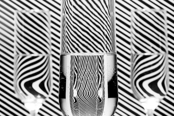 Black and white image of three filled champagne glass with water in front of a background with a diagonal line pattern, with a mirror reflection in the liquid