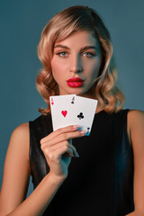 Blonde girl in black stylish dress showing two playing cards, posing against blue background. Gambling entertainment, poker, casino. Close-up.