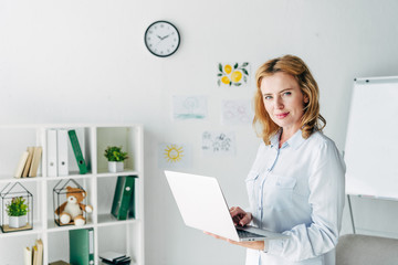 attractive child psychologist in shirt looking at camera and holding laptop