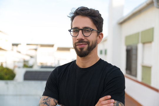 Joyful stylish guy in glasses posing on apartment balcony or terrace. Young man in casual standing outside with arms crossed, looking at camera and smiling. Male portrait concept