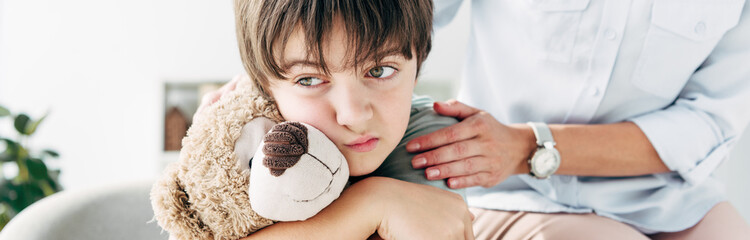 panoramic shot of kid with dyslexia holding teddy bear and child psychologist hugging him