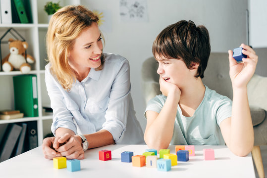 Smiling child psychologist looking at kid with dyslexia and sitting at table with building blocks