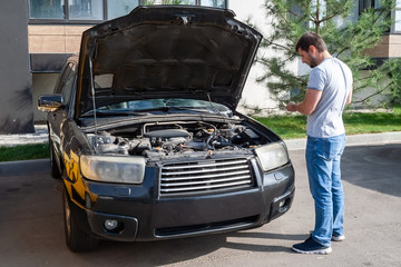 A thoughtful bearded man stands in front of a black car with an open hood during a repair....