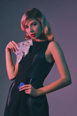 Blonde girl in black stylish dress holding some money and chips, posing against colorful background. Gambling entertainment, poker, casino. Close-up.