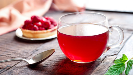 Delicious tartlet with raspberries, a cup of tea, a spoon and green raspberry leaf on an old wooden table near the window in backlight. 