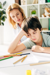 selective focus of sad kid with dyslexia sitting at table and child psychologist looking at him on background