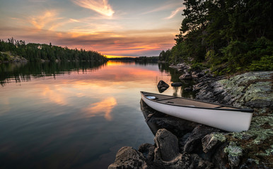 A lone canoe sits on shore on a peaceful lake at sunset. The odd colored clouds in the background are from a forest fire in Northwest Ontario, Canada in the summer of 2018.