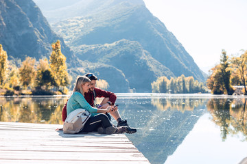 Two travel hikers using mobile phone while sitting in front of the lake in mountain.