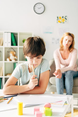 selective focus of thoughtful kid with dyslexia holding pencil and child psychologist on background - obrazy, fototapety, plakaty