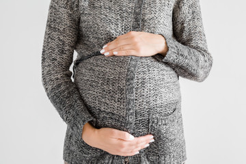 Young pregnant woman in knitted dark jacket touching big belly with hands. Isolated on gray...