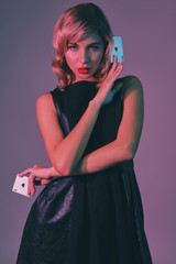 Blonde girl in black stylish dress showing two playing cards, posing against colorful background. Gambling entertainment, poker, casino. Close-up.