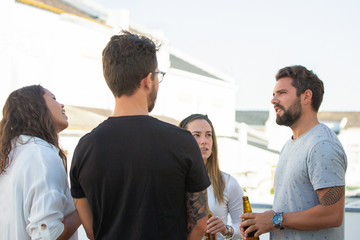 Group of friends sharing news over bottle of beer on outdoor terrace. Young men and women in casual meeting outside. Friendly hangout concept