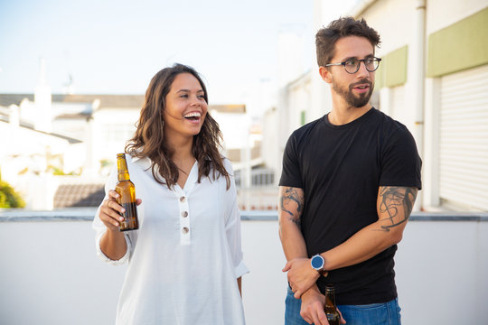 Happy couple talking, laughing, looking away and drinking beer on outdoor terrace. Young man and women in casual meeting outside. Hangout on rooftop concept