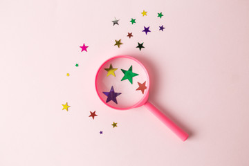 Top view of magnifier and different sizes multicolored stars on pastel rose background abstract.