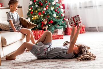 girl unwraps a gift while lying on a carpet opposite a Christmas tree in a house