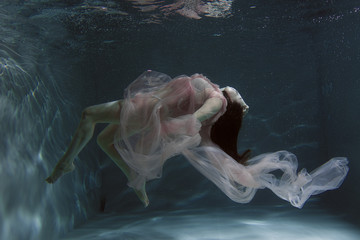 A girl with long dark hair swims underwater in a pink dress and with a crown on her head, like an...