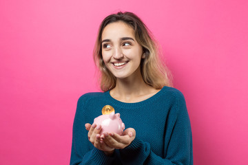 Fototapeta na wymiar Girl puts in the piggy bank physical Bitcoin.Young girl over pink background holding piggy bank and rejoices gesturing. The concept of reliability of cash investments and insurance.