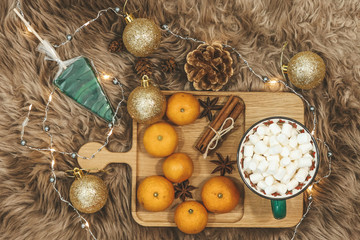 Wooden board with tangerines and cocoa with marshmallows, golden balls, cones, cinnamon and anice star, caramel on a background of brown fur. New Year flat lay