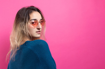 Romantic blonde-haired girl in trendy heart glasses posing with shy smile. Studio close-up portrait on a pink background.