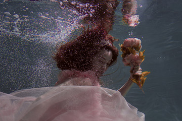 A girl with long dark hair swims underwater in a pink dress and with a crown on her head, like an...