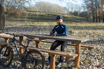 Cyclist in pants and fleece jacket on a modern carbon hardtail bike with an air suspension fork rides off-road. The guy is resting on a bench in the autumn park.