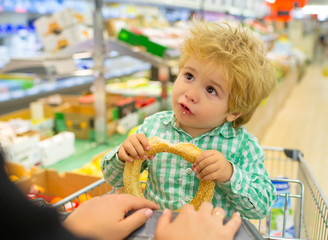 Cute baby in a shopping cart. Beautiful boy nibbles a bun while mom is shopping. Healthy Baby Food.