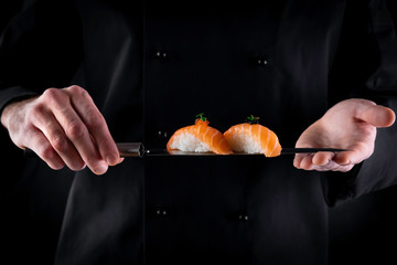 Sushi served on japanese knife in chef hands on dark background. Decorated salmon sashimi nigiri....