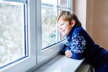 Happy adorable kid boy sitting near window and looking outside on snow on Christmas day or morning. Smiling healthy child fascinated observing snowfall and big snowflakes