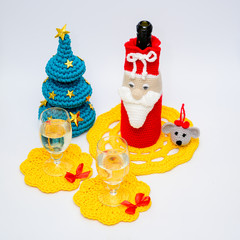 Home decor concept for new year and christmas. Photo of knitted decor thread for a bottle of champagne and a glass with a toy toy mouse, Christmas tree and yellow napkins on a white background.
