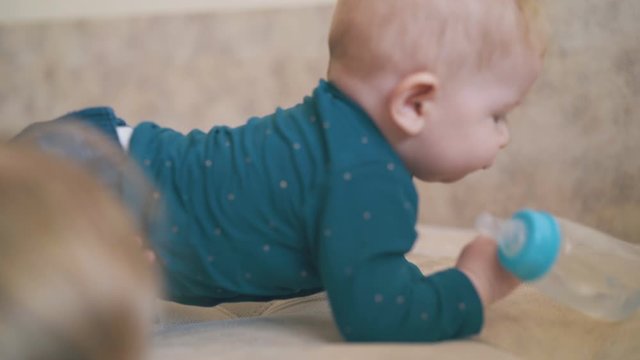 blonde little girl and funny baby trying to drink water from bottle on large beige bed in children room closeup