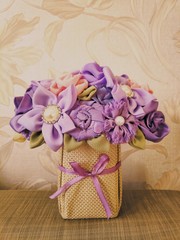photograph of a delicate bouquet of flowers from fabric of lilac and pink color in a basket of beige fabric with a purple bow on the background of cloth and plants