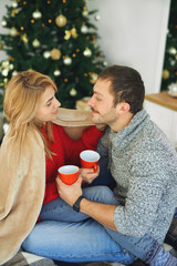 Romantic couple in love covered with warm soft blanket, embracing and holding cups with coffee at home with christmas tree on background.