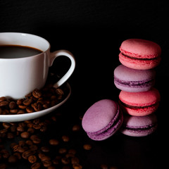 A white cup with black fragrant coffee stands on a black background with coffee zurnes next to colorful french cookies macaroons